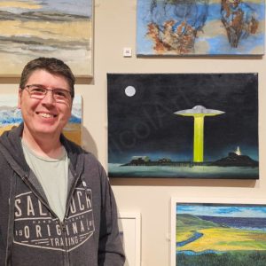 A photograph of David Nicol standing beside his painting of a UFO beaming up people from Mumbles Pier on a moonlit night. The painting is titled "A Souvenir from the Pier".